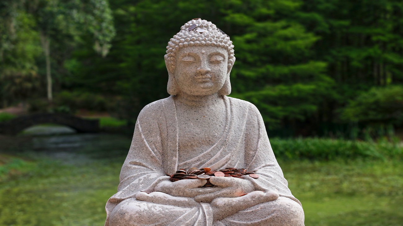 Foto de uma estátua de Buddha, segurando moedas sobre seu colo, representando a economia budista. No fundo, algumas árvores.