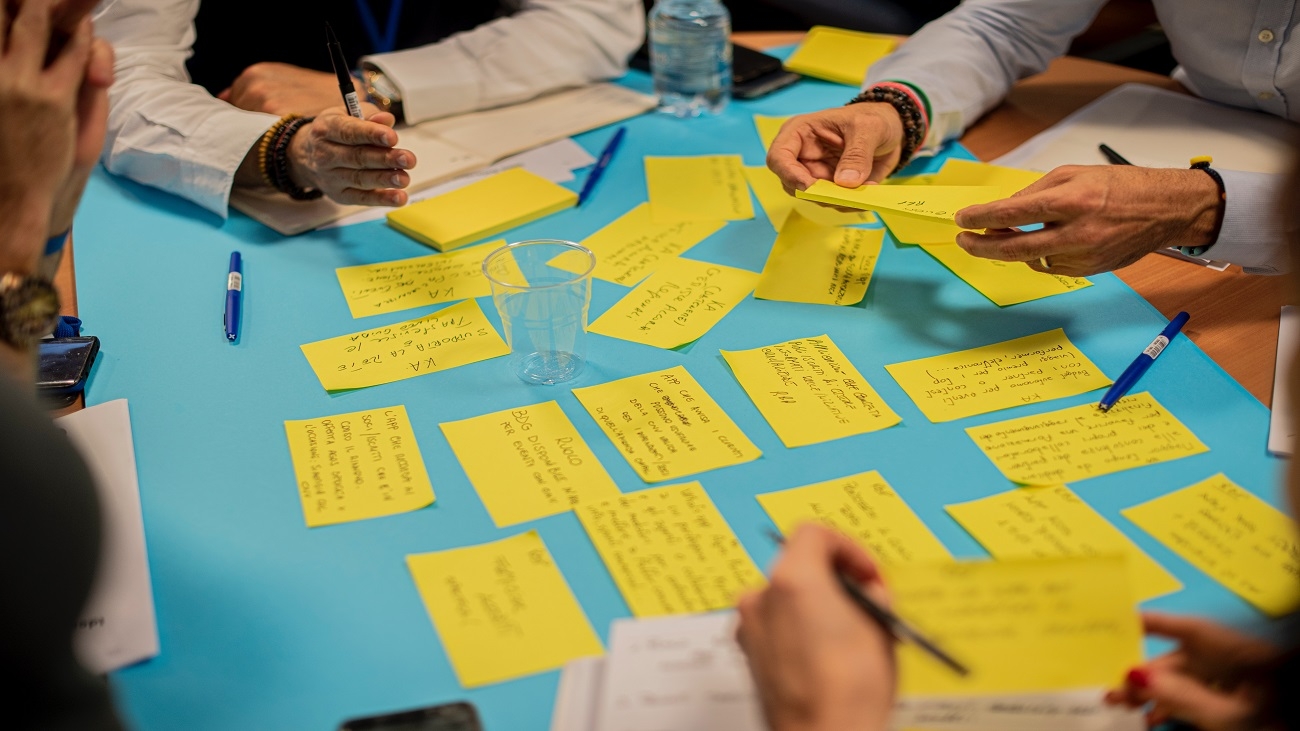 Foto de uma mesa com vários post its em cima, junto com algumas mão escrevendo em outros papeis.