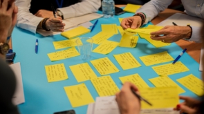 Foto de uma mesa com vários post its em cima, junto com algumas mão escrevendo em outros papeis.