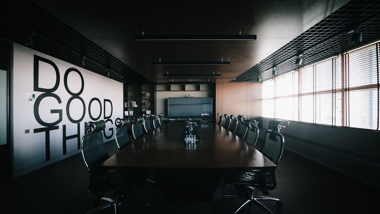 Uma sala de reuniões vazia, composta por uma grande mesa e diversas cadeiras ao seu redor. Na parede esquerda está escrito faça boas coisas.